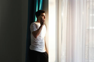 Handsome brunette bearded man stands before bright window and talks on his phone