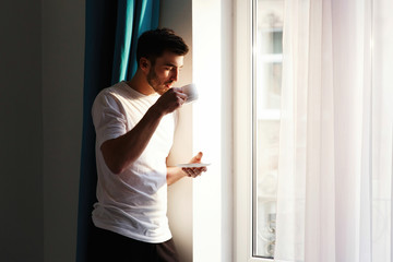 Fototapeta na wymiar Handsome brunette bearded man stands before bright window and drinks coffee