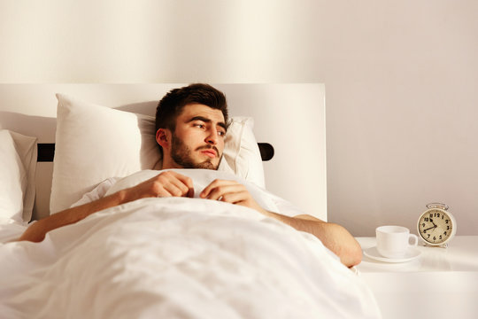 Brunette man looks tired lying in the bed with clock on bedside chest