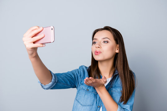 Close up portrait of sexy businesswoman, student, freelancer making selfie on her smartphone and blowing kiss to the front camera while standing over grey background