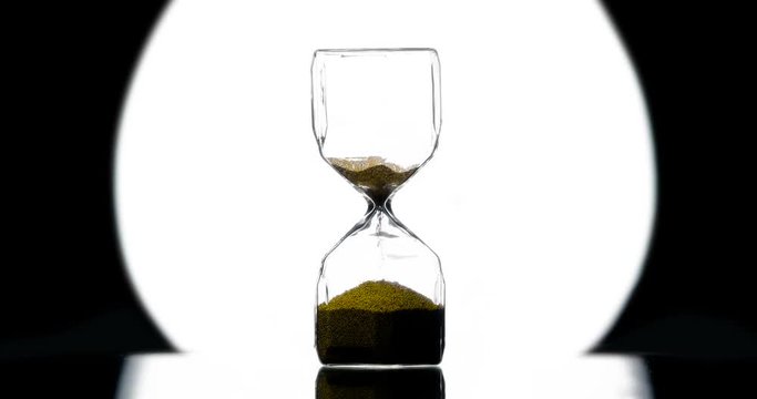 Deadline. Hourglass in Style Noir.Golden sand is strewed against the background of a bright round beam of a spotlight on a black. Filmed at a speed of 120fps