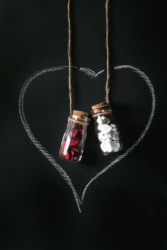 Two bottles full of white and red decorative hearts inside drawn by chalk heart shape over black background. St Valentine concept. Copy space