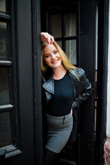 Blonde fashionable girl in long black leather coat posed against wooden door at old house.