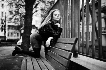 Blonde fashionable girl in long black leather coat posed on bench.