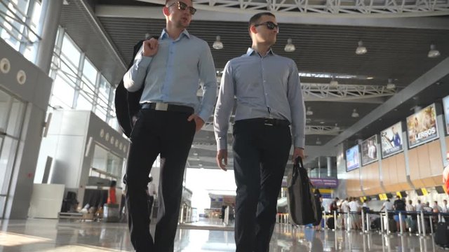 Two young businessmen walking in terminal at airport and talking. Men stepping at business center and speaking. Front view. Slow motion