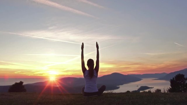 Young attractive girl sitting on the peak of a mountain. Practicing yoga. Surya Namaskar or Salutation to the Sun