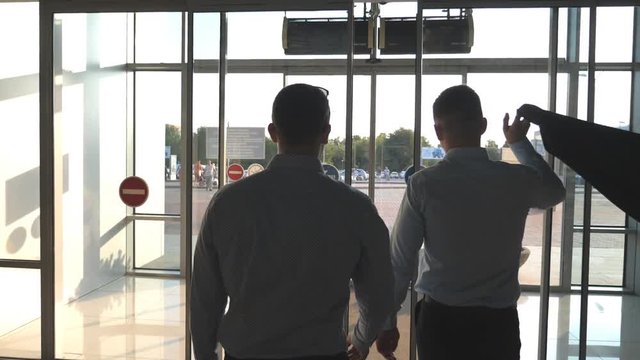 Two businessmen walking from the airport. Two man going through glass automatic door to the street. Sun flare at background. Trip or travel concept. Back view Slow motion