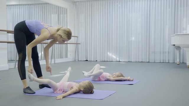The ballerina helps two girls to stretch the legs during the ballet class. The little dancers in pink dresses lie on the back on the mats with spreaded legs and the teacher carefully pushes the feets