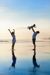 Father tossing high in air baby son, mother jumping by water pool. Happy family walk with fun by sunset black sand beach with sea surf. Active parents, outdoor activity on summer vacation with kids.