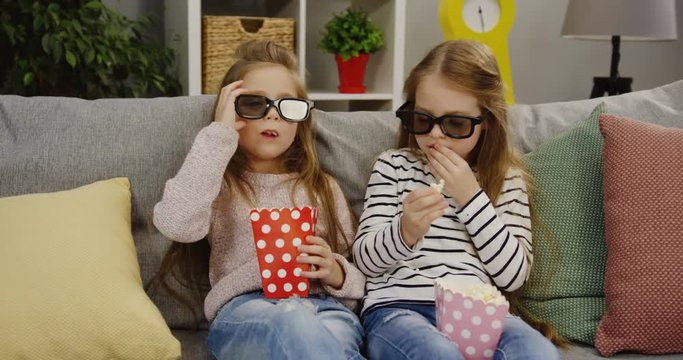 Two funny pretty little girls with long hair and wearing glasses sitting on the sofa with pillows and eating popcorn. Cute friends. Comfortable living room. Indoor