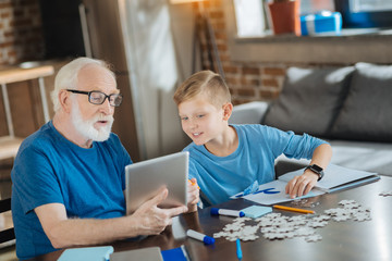 Digital device. Joyful nice happy boy pointing at the tablet and smiling while helping his grandfather to use it