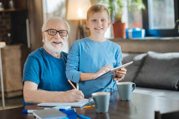 Modern igen. Nice cheerful delighted boy having a tablet in his hands and smiling while standing together with grandfather