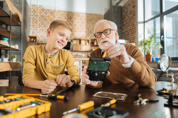 Be attentive. Smart nice skillful man holding a broken electronic device and showing his grandson what to do while fixing it