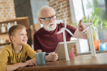 Sharing knowledge. Nice wise elderly man pointing at the windmill model and talking to his grandson while sharing his knowledge