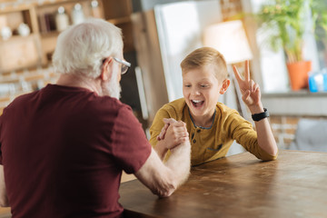 Who is stronger. Happy cheerful nice grandfather and grandson looking at each other and smiling while doing arm wrestling