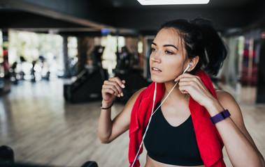 Fototapeta na wymiar Horizontal portrait young Caucasian woman runner corrects headphones while running in the gym, wearing black sportswear and red towel on neck. People, sport and fitness concept.