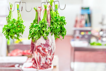 Whole raw lamb leg hanged on hook with bunch of parsley at market or shop. Meat prepared for cook