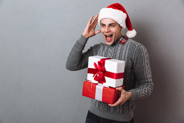 Pleased screaming man in sweater and christmas hat holding gifts
