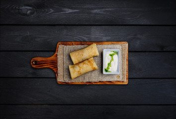 Thin stuffed pancakes or russian blini with sour cream. On a wooden board. Top view. Black wooden...