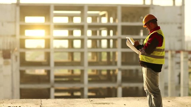 Master builder looks at new construction site and makes notes in works documentation. Worker in hard hat stands on project site and looks at new building structure, beautiful sunset at background