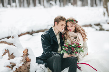 Couple in love with a bouquet are sitting on the log on background of the snowy forest. Winter wedding. Artwork - 182723810
