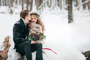 Groom is kissing his bride on the temple on background of the snowy forest. Winter wedding. Artwork