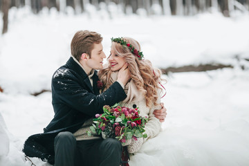 Couple in love with a bouquet are sitting on the log on background of the snowy forest. Winter wedding. Artwork - 182723870