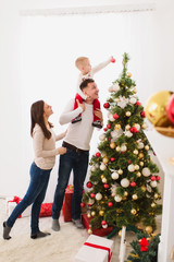 Happy young cheerful parents with cute little son. Child boy on daddy shoulders decorating New Year tree with toy ball in light room at home. Christmas good mood. Family, love and holiday 2018 concept