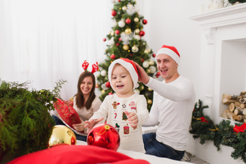 Happy young parents with cute little son in hat. Child boy with big red tree toy ball standing in decorated New Year light room at home. Christmas good mood. Family, love and holiday 2018 concept.