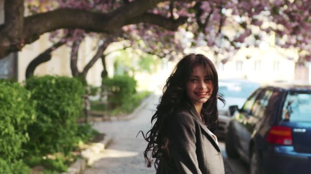 Back view of long-haired brunette walking down the alley, then she whirls and smiles right towards the camera. Positive emotions, playful mood, seductiveness.