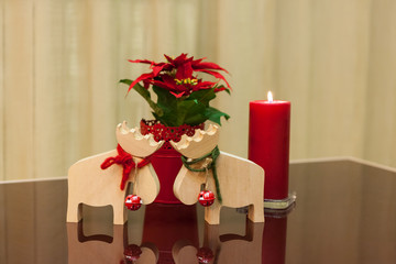 wooden decorative interior Christmas toys moose deer with red and green ropes tied around the neck and red bells with a lit red candle and with poinsettia in a red pot on a glass table