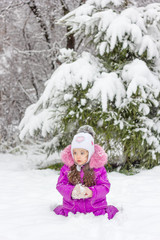 Little kid girl sitting near pine tree covered by snow on winter forest.