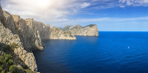 Fototapeta na wymiar A panoramic picture of the cliffs on the Pollença coastline and a single white sailboat sailing on the calm sea on a nice sunny day in Mallorca