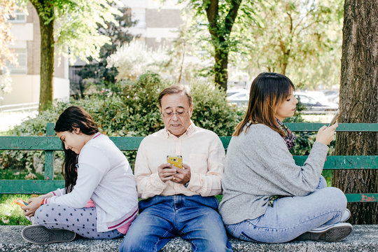 .A father with his daughters sitting on a stone bench, using their mobile phone without looking at each other and no talking. Ingnoring themselves. Lifestyle portrait...