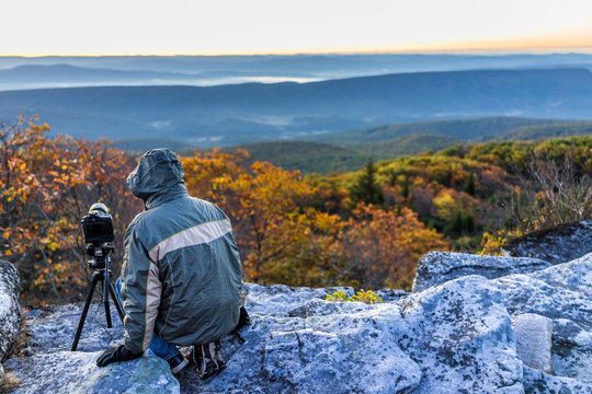 Sitting young man photographer with camera, gear, and tripod on cold autumn morning in jacket in Bear Rocks, West Virginia taking pictures of sunrise