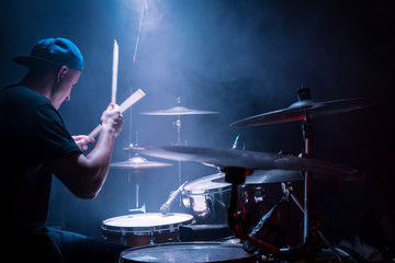 Drummer in a cap and headphones plays drums at a concert under blue light in a smoke
