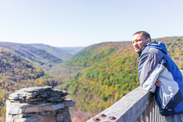 Fototapeta na wymiar Young man in blue jacket looking at canaan valley mountains in Blackwater falls state park in West Virginia during colorful autumn fall season with yellow foliage on trees, rock cliff at Lindy Point