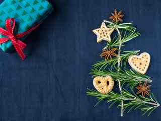 Christmas tree made from rosemary, anise star and gingerbread cookies.  Christmas box with red ribbon on blue background.