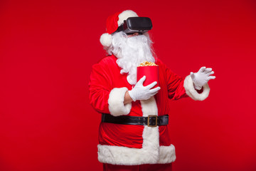 Santa Claus wearing virtual reality goggles and a red bucket with popcorn, on a red background. Christmas