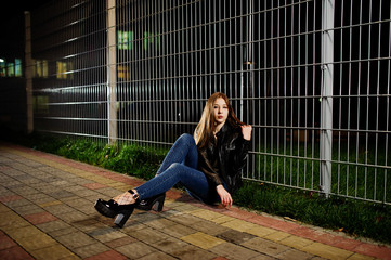 Night portrait of girl model wear on jeans and leather jacket against iron fence.