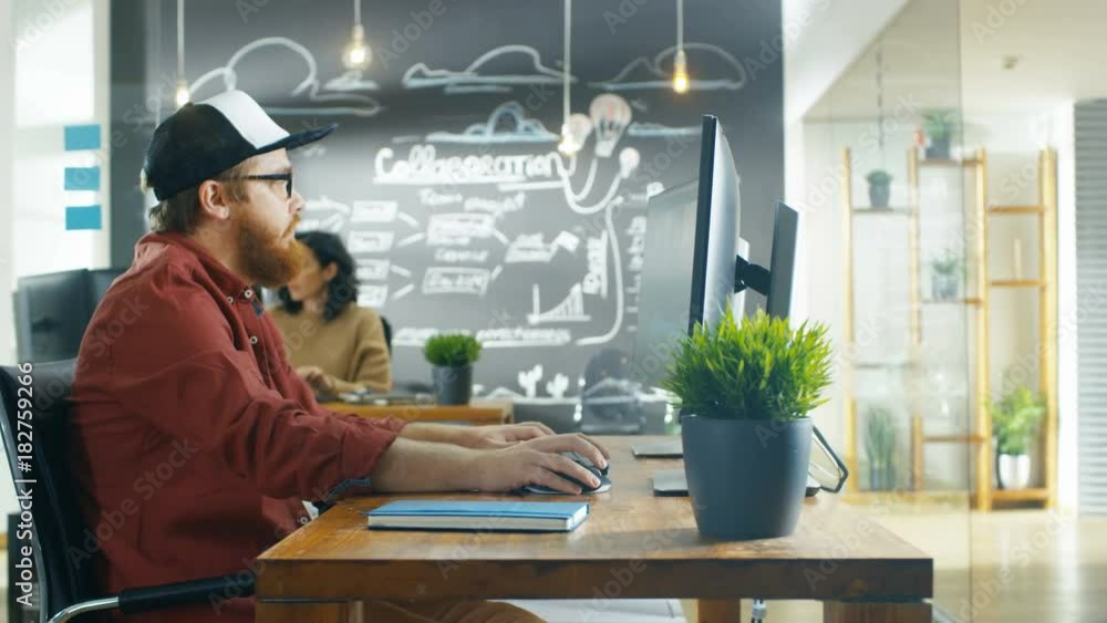 Poster in the creative agency videographer in a cap works on personal computer with two monitors, he's doin