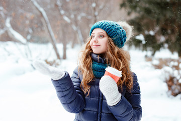 Winter walk in the park or the wood. The young beautiful girl the blonde with blue eyes in a warm cap and a down-padded coat catches a hand in a snowflake mitten. There is snowfall.