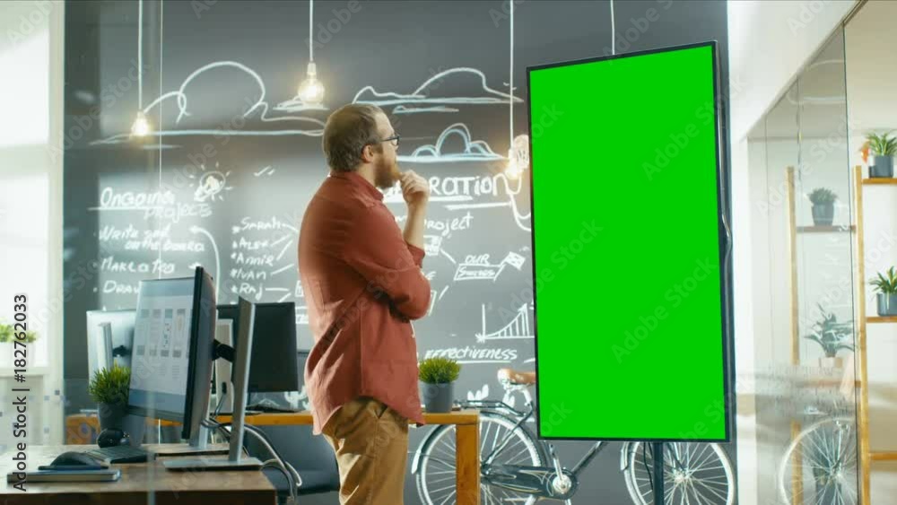 Poster Male App Developer Looks at Interactive Whiteboard with Mock-up Green Screen. He's Thinking on a Problem Solution. In the Background Creative Office with Chalkboard. 