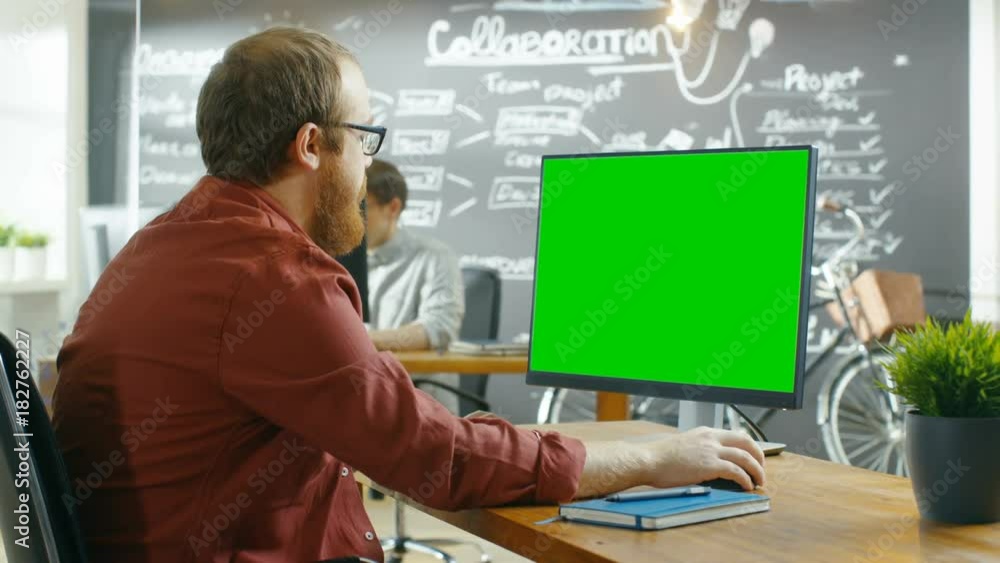 Poster Man Works on the Personal Computer with Green Mock-up Screen. In the background Creative Office with Chalkboard Wall. Shot on RED EPIC-W 8K Helium Cinema Camera.