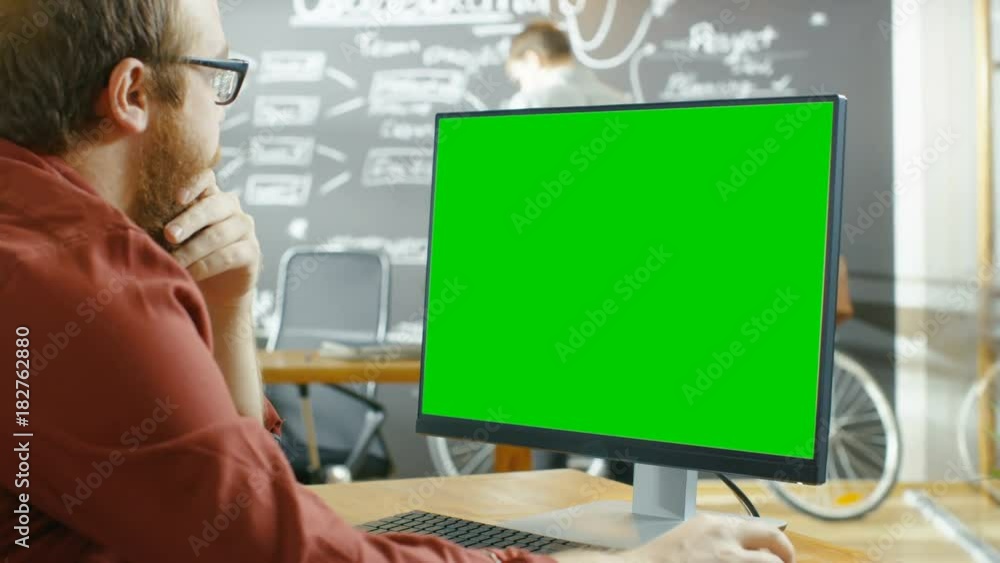 Aufkleber Man Works on the Personal Computer with Green Mock-up Screen. In the background Creative Office with Man Drawing on Chalkboard Wall. Shot on RED EPIC-W 8K Helium Cinema Camera.