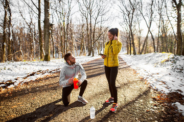 Young sporty happy couple in sportswear making a break with water and music after jogging through the forest in the sunny winter morning.