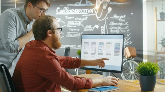 Two Male Mobile Game Application Developers Talk while Working on a Personal Computer, Designing Wireframe. Coworkers Work in the Creative Indie Studio. Shot on RED EPIC-W 8K Helium Cinema Camera.