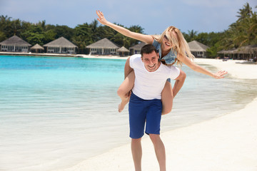 Portrait of smiling man carrying woman on his back along the sea shore. Guy giving piggyback ride...