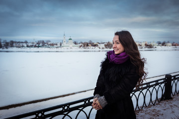 Young woman with beautiful long hair in purple scarf standing near the snowy river at the city promenade and looking away. Cold day, Cloudy sky