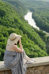 Girl sits on hill and takes pictures against the background of a forest and river